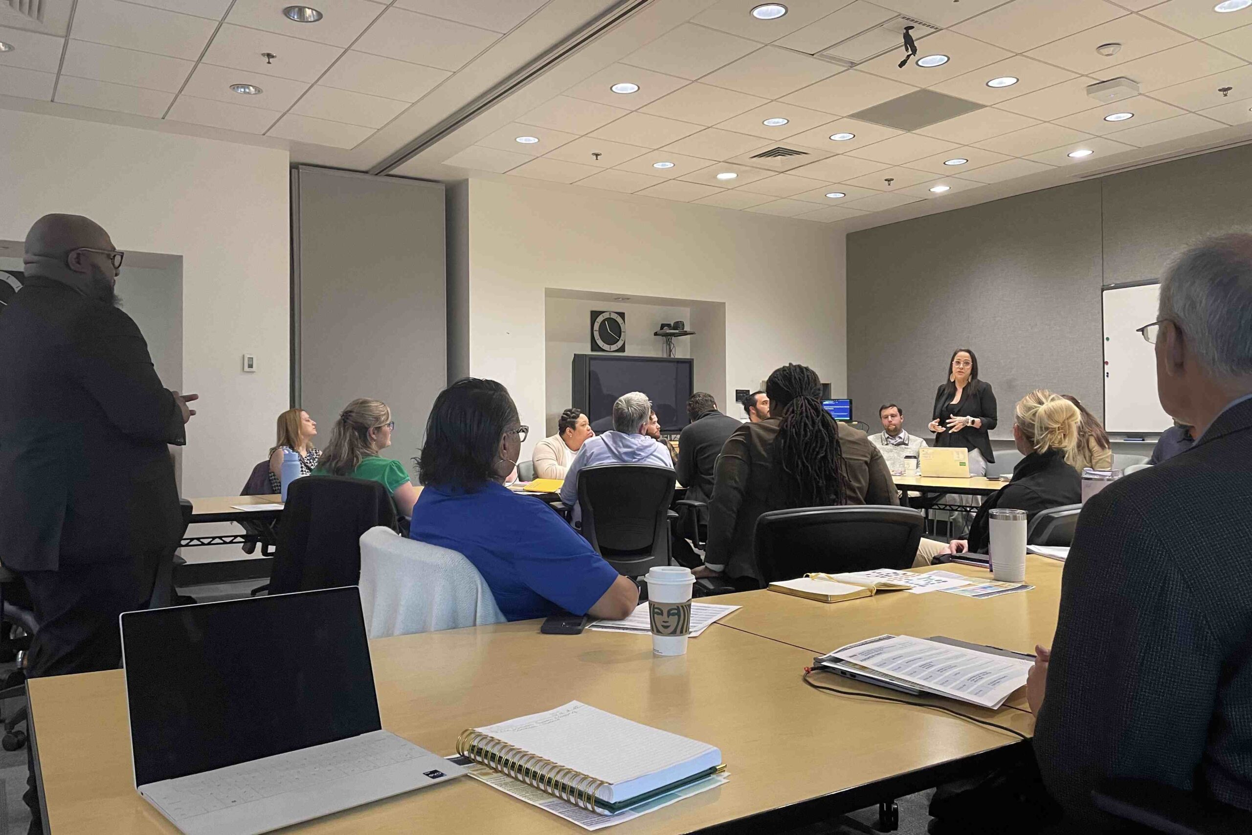 A group of people sitting in a conference room listening to a women standing at the front of the room.